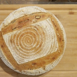May include: A round loaf of sourdough bread with a swirl pattern on the top crust. The bread is on a wooden chopping board.