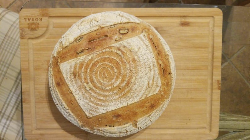 May include: A round loaf of sourdough bread with a swirl pattern on the top crust. The bread is on a wooden chopping board.
