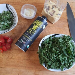 May include: A wooden cutting board with a variety of ingredients for a salad, including fresh basil, cherry tomatoes, chopped kale, a bottle of extra virgin olive oil, a container of garlic, and a chef's knife.