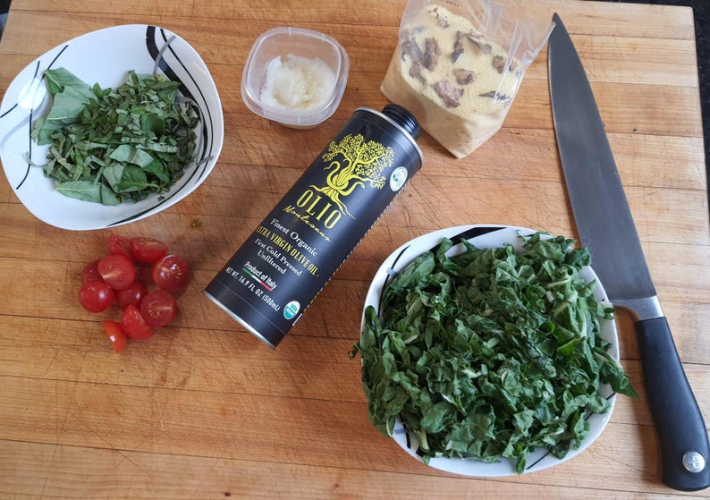 May include: A wooden cutting board with a variety of ingredients for a salad, including fresh basil, cherry tomatoes, chopped kale, a bottle of extra virgin olive oil, a container of garlic, and a chef's knife.