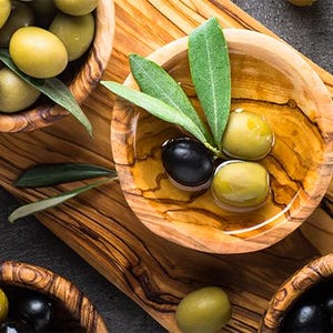 May include: A close-up of three wooden bowls filled with green olives and black olives. The bowls are on a wooden cutting board with olive branches and leaves.