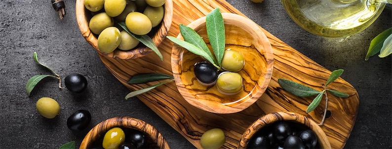May include: A close-up of three wooden bowls filled with green olives and black olives. The bowls are on a wooden cutting board with olive branches and leaves.