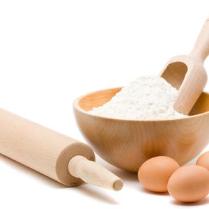 May include: A wooden bowl filled with white flour, a wooden scoop, a wooden rolling pin, and three brown eggs on a white background.