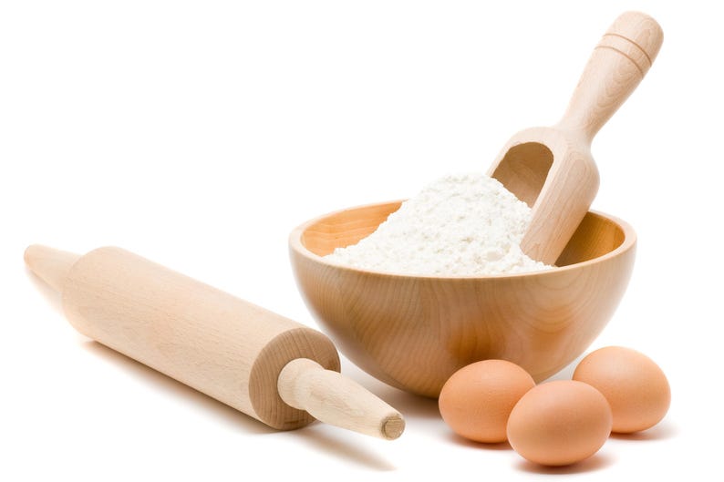 May include: A wooden bowl filled with white flour, a wooden scoop, a wooden rolling pin, and three brown eggs on a white background.