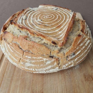 May include: A round loaf of sourdough bread with a spiral pattern on top. The bread is on a wooden chopping board.