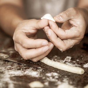 May include: Close-up of a person's hands shaping dough into small, white dumplings. The dough is on a wooden surface covered in flour.