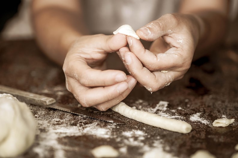 May include: Close-up of a person's hands shaping dough into small, white dumplings. The dough is on a wooden surface covered in flour.