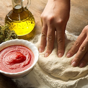 May include: A person's hands kneading a ball of dough on a floured surface. There is a bowl of tomato sauce, a bowl of shredded cheese, and a bottle of olive oil in the background.