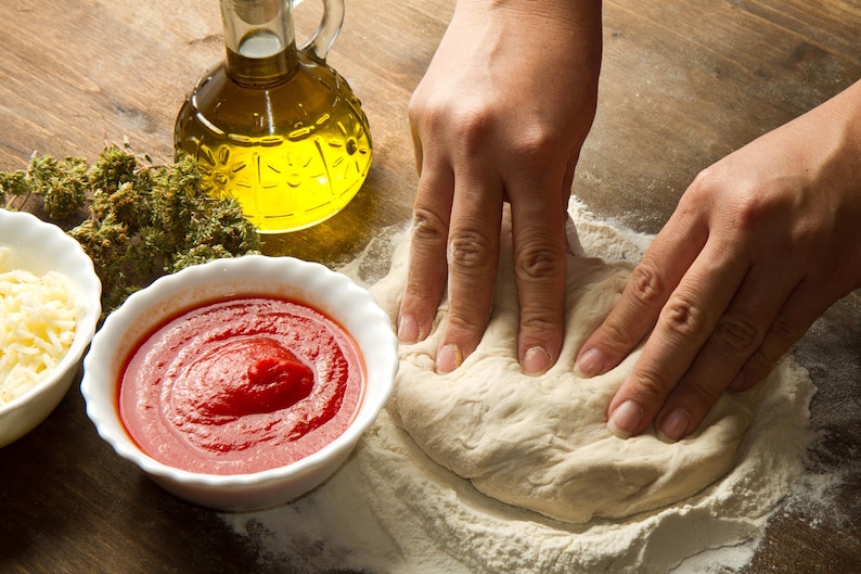 May include: A person's hands kneading a ball of dough on a floured surface. There is a bowl of tomato sauce, a bowl of shredded cheese, and a bottle of olive oil in the background.