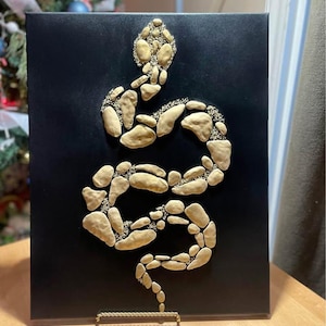 Puede incluir: Un lienzo negro con un diseño de serpiente dorada hecho de pequeñas piezas redondeadas. La serpiente está enroscada y tiene la cabeza levantada. El lienzo está sobre un soporte dorado.