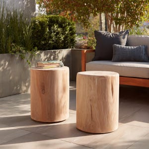 May include: Two light brown wooden side tables with a cylindrical shape. One table has a book on top. The tables are outdoors on a tiled patio, with a couch and greenery in the background. The tables are approximately 18 inches in height.