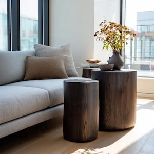May include: Two dark brown wooden side tables with a cylindrical shape. The tables are placed in front of a light gray sofa and a window. A vase with dried flowers sits on one table. The tables are made of wood.