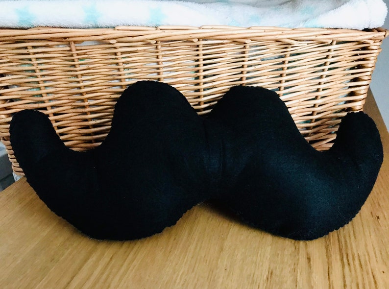 Pu&ograve; includere: Un cuscino a forma di baffi in feltro nero. Il cuscino &egrave; appoggiato su una superficie di legno.