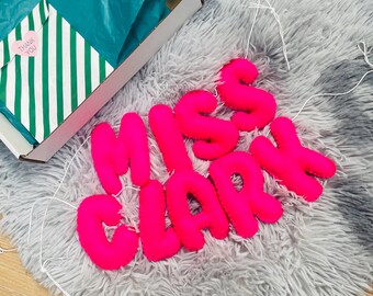 May include: Pink felt letters spelling "Miss Clark" on a gray fluffy rug. The letters are connected with white string.
