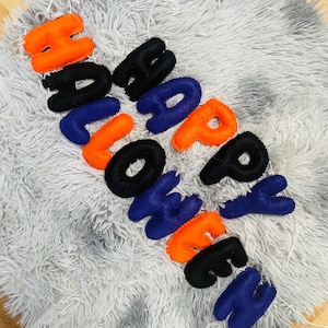 May include: A felt banner spelling "HALLOWEEN" in orange, black, and blue letters. The letters are strung together with white string and rest on a fluffy, gray surface. A festive decoration for Halloween.