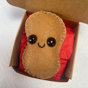 Peut inclure: Un jouet en peluche en forme d'arachide en feutre marron avec un visage souriant et des yeux noirs, assis dans une boîte en carton avec du papier de soie rouge.