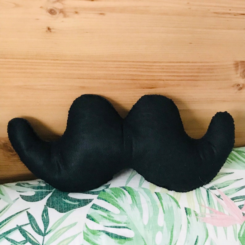 Pu&ograve; includere: Un cuscino di stoffa nero a forma di baffi. Il cuscino &egrave; a forma di baffi con due estremit&agrave; arrotondate grandi e una sezione centrale pi&ugrave; piccola. Il cuscino &egrave; su una superficie con motivo verde e bianco.