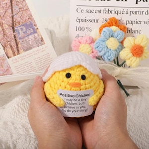 Op de afbeelding: Een handgemaakte gele gehaakte kuiken met een witte eierschaal top, zwarte ogen en een oranje snavel. De kuiken houdt een bord vast met de tekst "Positive Chicken". Een boeket kleurrijke gehaakte bloemen staat op de achtergrond.