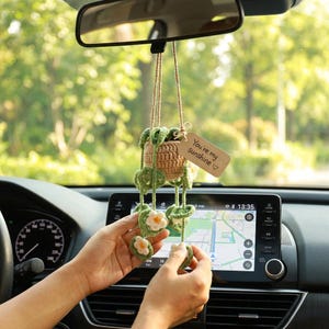May include: A crocheted car charm featuring a hanging basket with green leaves and white flowers. The charm is suspended by a string and includes a tag that reads "You're my sunshine".