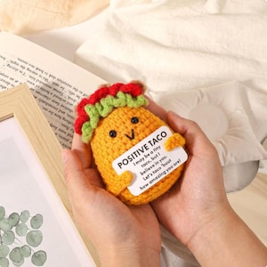 Może przedstawiać: Szydełkowe taco z uśmiechniętą twarzą i czerwonymi, zielonymi i brązowymi wykończeniami. Taco trzyma biały znak z napisem "POSITIVE TACO" z inspirującą wiadomością. Taco jest trzymane w dwóch rękach.