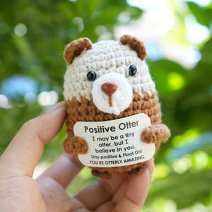 May include: A crocheted otter figure in brown and cream, holding a sign with the words "Positive Otter" and an inspirational message. The otter has black eyes and a brown nose. A handmade item.