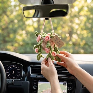 Könnte beinhalten: Gehäkelter Autoanhänger mit rosa und grünen Blumen in einem hellrosa Topf, der am Rückspiegel eines Autos hängt. Der Anhänger hat ein Etikett mit der Aufschrift "You are so loved" mit einem Herzsymbol.