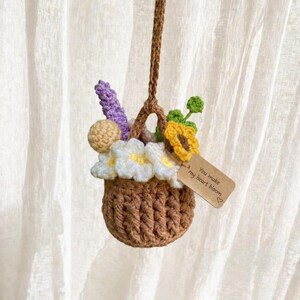 May include: A crocheted brown basket filled with colourful flowers, including white daisies, a yellow sunflower, and purple lavender. A small tag reads "You make my heart bloom". The basket hangs from a brown cord.