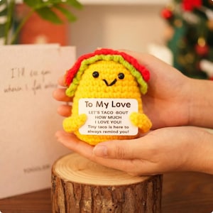May include: A handmade yellow crochet taco with a red and green shell. The taco has a smiling face and holds a sign that reads "To My Love" and a message about love. The taco is displayed on a wooden slice.
