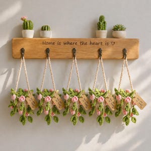 Puede incluir: Una estantería de madera con ganchos exhibe cinco cestas de flores de ganchillo colgantes. Cada cesta presenta rosas rosas y hojas verdes, con una etiqueta que dice "You are so loved". La estantería también contiene pequeños cactus y suculentas en macetas. El texto "Home is where the heart is" está inscrito en la estantería.