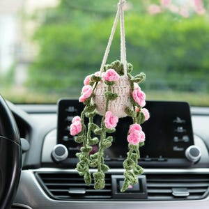 Op de afbeelding: Een hangende gehaakte plant met roze bloemen en groene wijnstokken. De plant staat in een beige pot en wordt opgehangen met een wit koord. Het decoratieve item hangt in een auto.