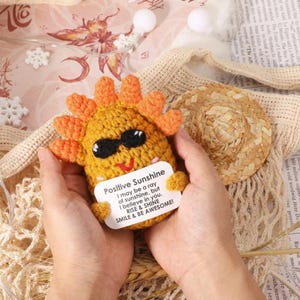 Op de afbeelding: Een gehaakte gele zon met oranje stralen en een zwarte zonnebril, met een witte kaart met de tekst "Positive Sunshine". De kaart luidt: "I may be a ray of sunshine, but I believe in you. RISE & SHINE SMILE & BE AWESOME!"