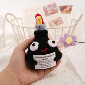 May include: A black crocheted "Emotional Support Bomb" with white eyes, a red smile, and a red and yellow top. A white tag reads "Feeling stressed? Squeeze tightly. Aim and release. Satisfaction guaranteed!"