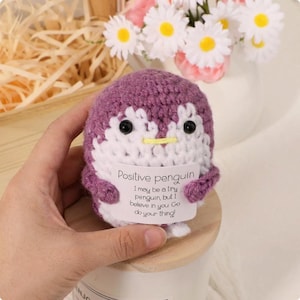Puede incluir: Un pingüino de ganchillo en morado y blanco, sosteniendo un letrero que dice "Positive penguin". El pingüino tiene ojos negros y un pico amarillo. El letrero dice "I may be a tiny penguin, but I believe in you. Go do your thing!"