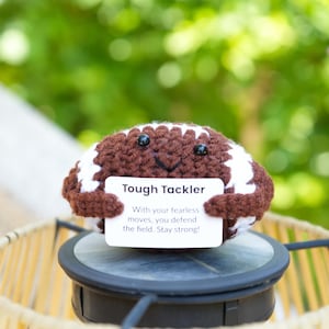 以下が含まれることがあります： 笑顔の顔をした、茶色と白の編み物フットボール。 「Tough Tackler」と書かれたサインを持っており、「With your fearless moves, you defend the field. Stay strong!」というテキストが含まれています。