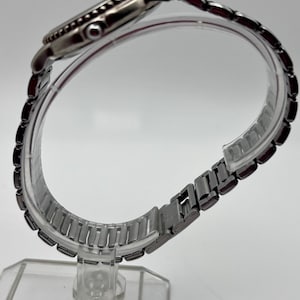 May include: A silver wristwatch with a black bezel and a metal link strap. The watch is presented on a clear plastic stand, highlighting its side view and strap design. The watch face is not visible.