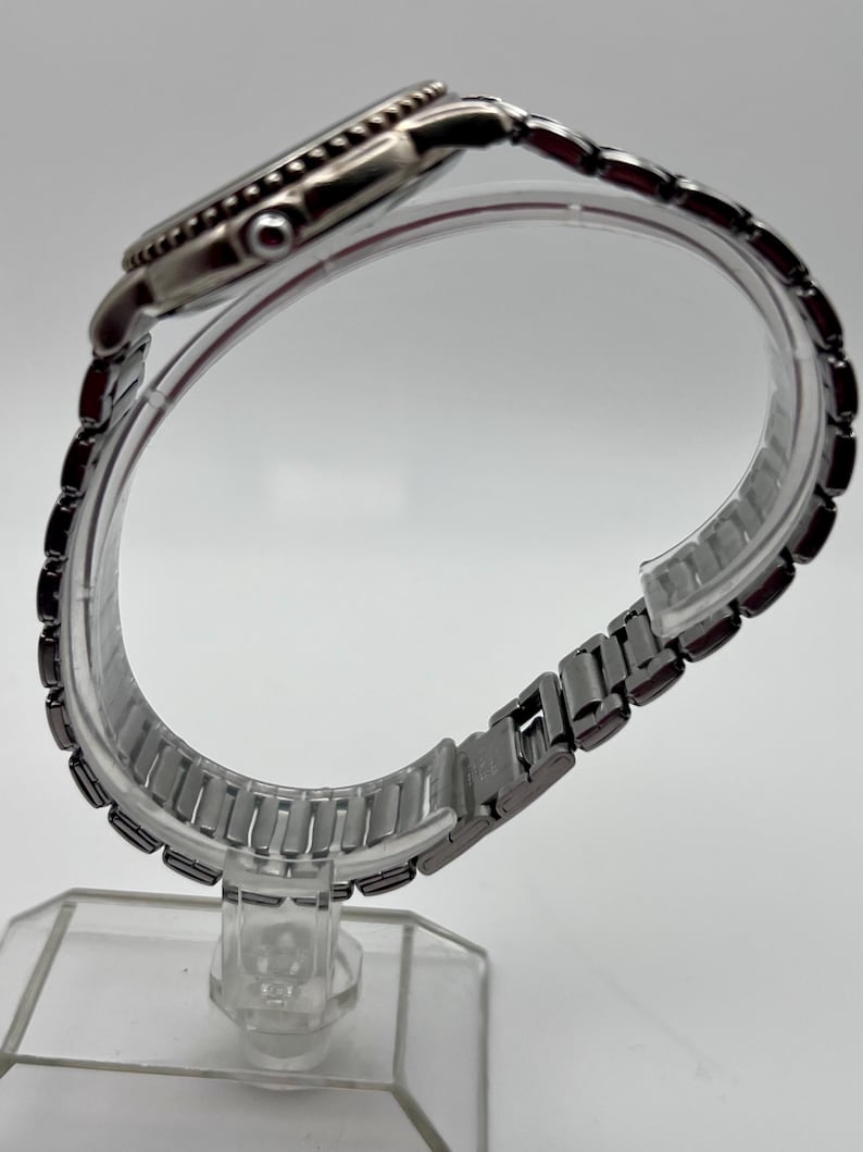May include: A silver wristwatch with a black bezel and a metal link strap. The watch is presented on a clear plastic stand, highlighting its side view and strap design. The watch face is not visible.