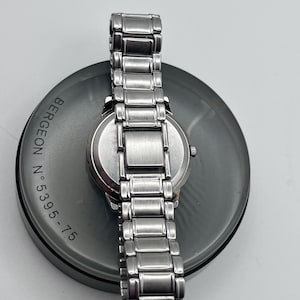 May include: A stainless steel wristwatch with a silver strap rests on a dark grey circular surface. The watch face is silver, and the strap is made of rectangular links. The surface has the text "BERGEON N.5395-75" printed on it.