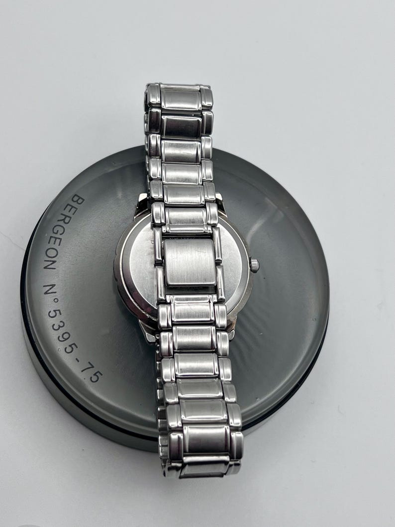 May include: A stainless steel wristwatch with a silver strap rests on a dark grey circular surface. The watch face is silver, and the strap is made of rectangular links. The surface has the text "BERGEON N.5395-75" printed on it.