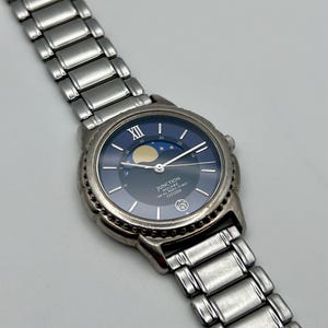May include: A silver wristwatch with a dark blue face, moon phase indicator, and silver hands. The watch has a stainless steel strap and the text "JUNCTION SQUARE FOR ALL TIME TO MEET CITIZEN" is printed on the face.