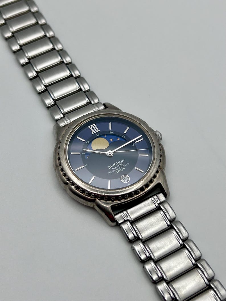 May include: A silver wristwatch with a dark blue face, moon phase indicator, and silver hands. The watch has a stainless steel strap and the text "JUNCTION SQUARE FOR ALL TIME TO MEET CITIZEN" is printed on the face.