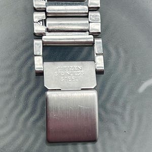 May include: A close-up of a silver Citizen stainless steel watch strap. The strap is made of rectangular links and has a clasp with the text "CITIZEN STAINLESS STEEL". The watch strap is on a reflective surface.