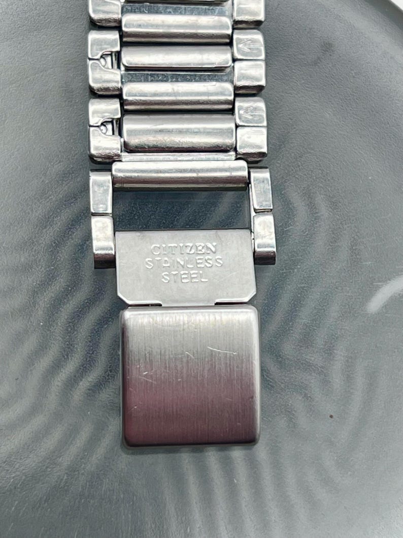 May include: A close-up of a silver Citizen stainless steel watch strap. The strap is made of rectangular links and has a clasp with the text "CITIZEN STAINLESS STEEL". The watch strap is on a reflective surface.