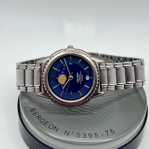 May include: A silver wristwatch with a dark blue face and a moon phase display. The watch has a stainless steel strap and a round bezel. It is presented on a grey and black stand, with the text "BERGEON" and "5395-75" visible.
