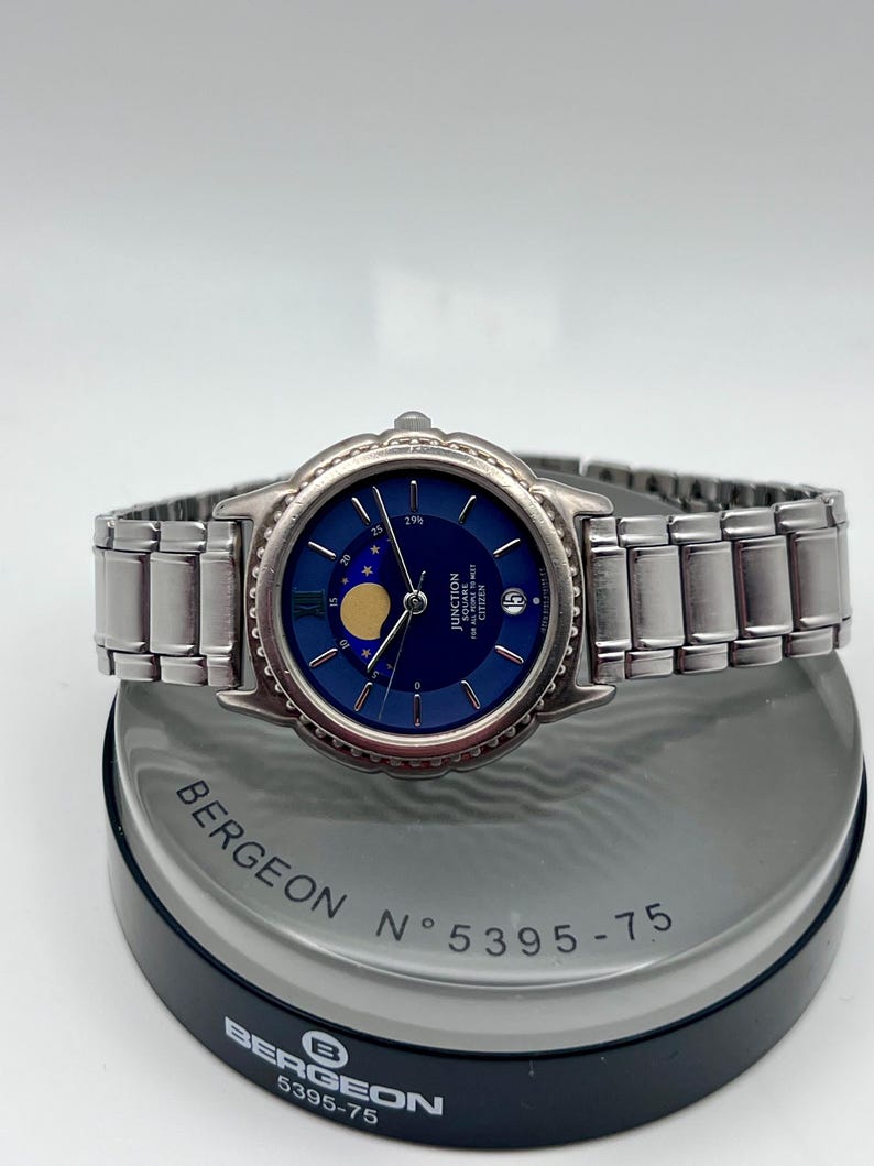 May include: A silver wristwatch with a dark blue face and a moon phase display. The watch has a stainless steel strap and a round bezel. It is presented on a grey and black stand, with the text "BERGEON" and "5395-75" visible.