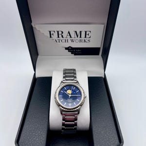 May include: A silver wristwatch with a blue face and a moon phase display, presented in a black box. The watch has a metal strap and is displayed on a white cushion. The box includes a card that reads "FRAME WATCH WORKS".