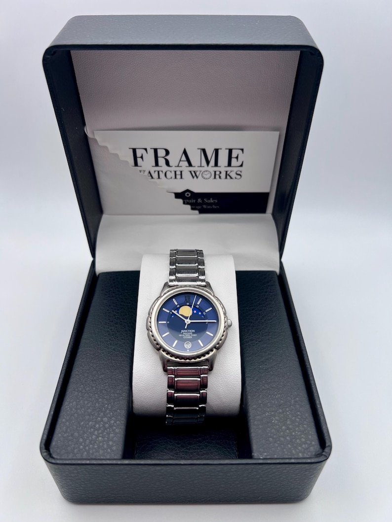 May include: A silver wristwatch with a blue face and a moon phase display, presented in a black box. The watch has a metal strap and is displayed on a white cushion. The box includes a card that reads "FRAME WATCH WORKS".