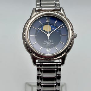 May include: A silver Citizen Junction Square watch with a dark blue face. It features a moon phase indicator and the text "JUNCTION SQUARE FOR ALL PEOPLE TO MEET CITIZEN". The watch has a silver band and a date display at the 3 o'clock position.