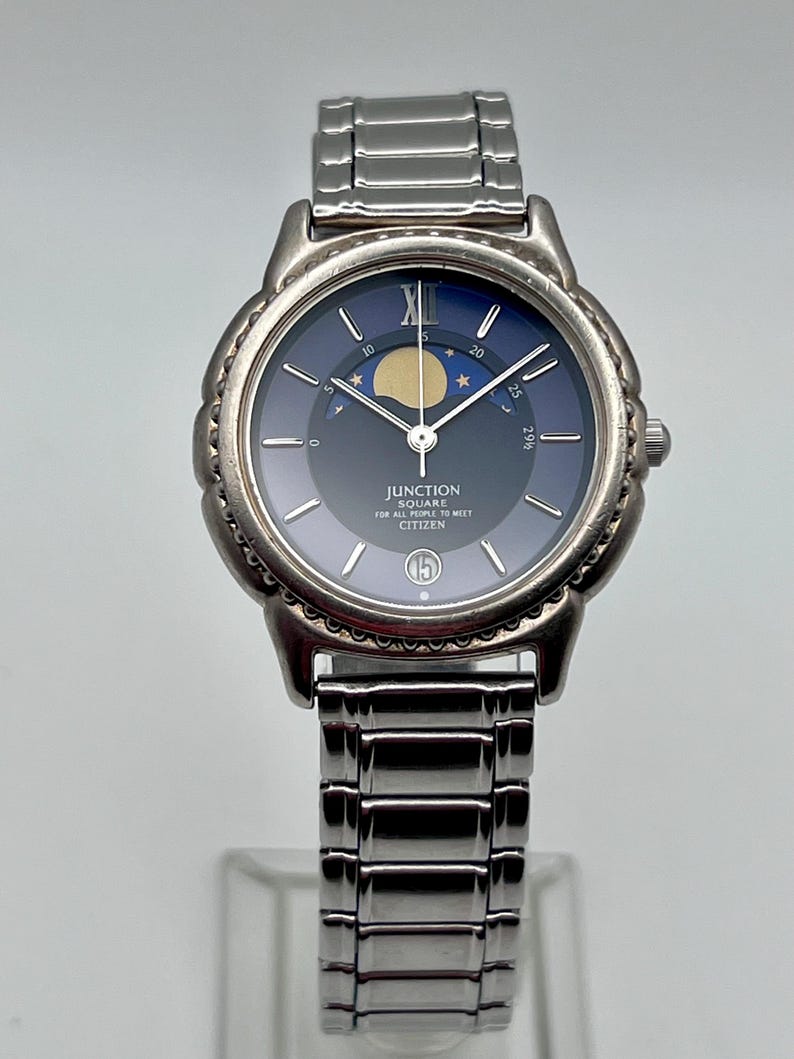 May include: A silver Citizen Junction Square watch with a dark blue face. It features a moon phase indicator and the text "JUNCTION SQUARE FOR ALL PEOPLE TO MEET CITIZEN". The watch has a silver band and a date display at the 3 o'clock position.