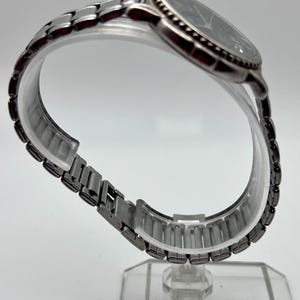 May include: A silver wristwatch with a dark blue face and a linked metal strap. The watch is displayed on a clear, curved stand, highlighting the strap's design. The watch face features silver hands and markers.