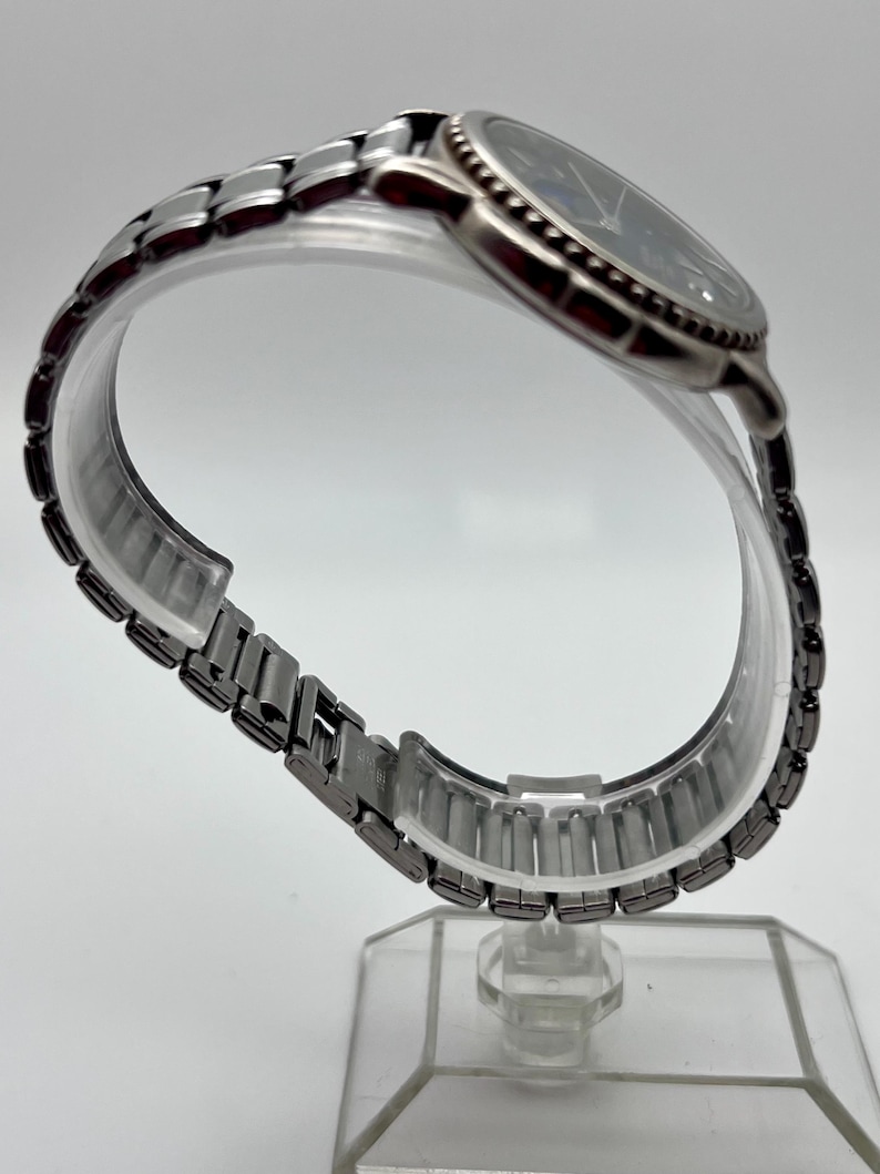 May include: A silver wristwatch with a dark blue face and a linked metal strap. The watch is displayed on a clear, curved stand, highlighting the strap's design. The watch face features silver hands and markers.